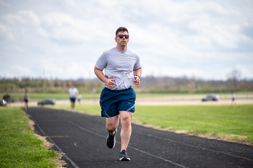 One man, wearing Air Force fitness attire, runs on a black track, another person can be seen out of focus in the distant background