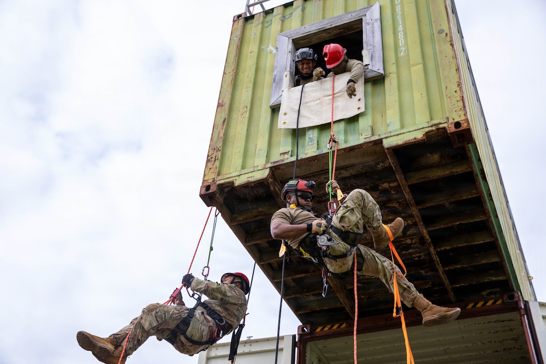 Two National Guardsmen wearing harnesses ascend toward a lifted storage container as fellow service members watch from above out of a window.