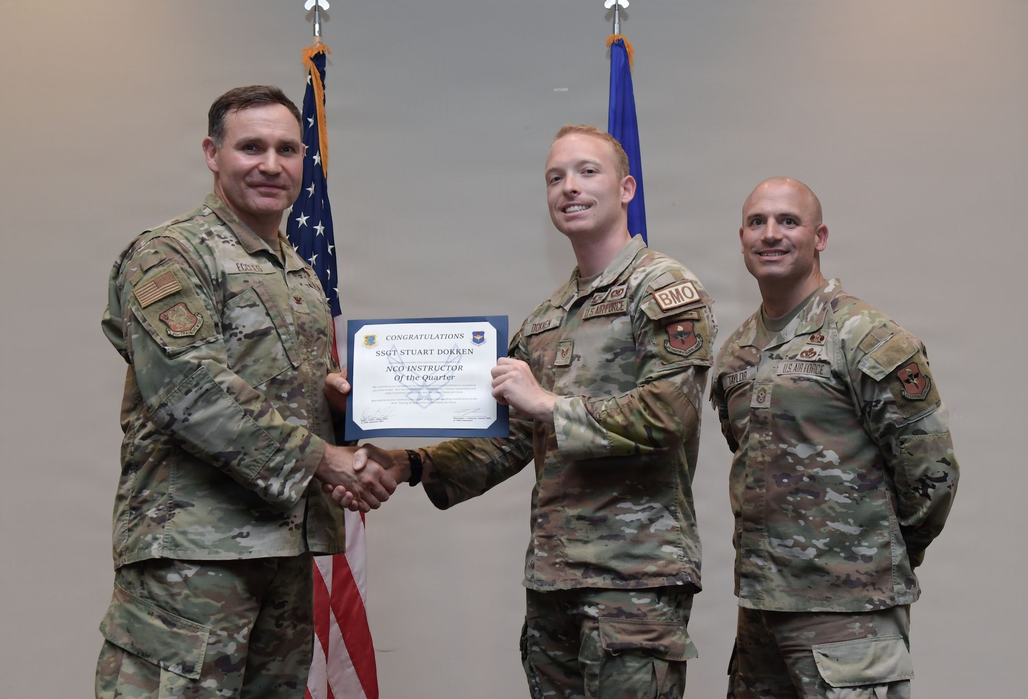 Three men in military uniform stand on stage holding a certificate.