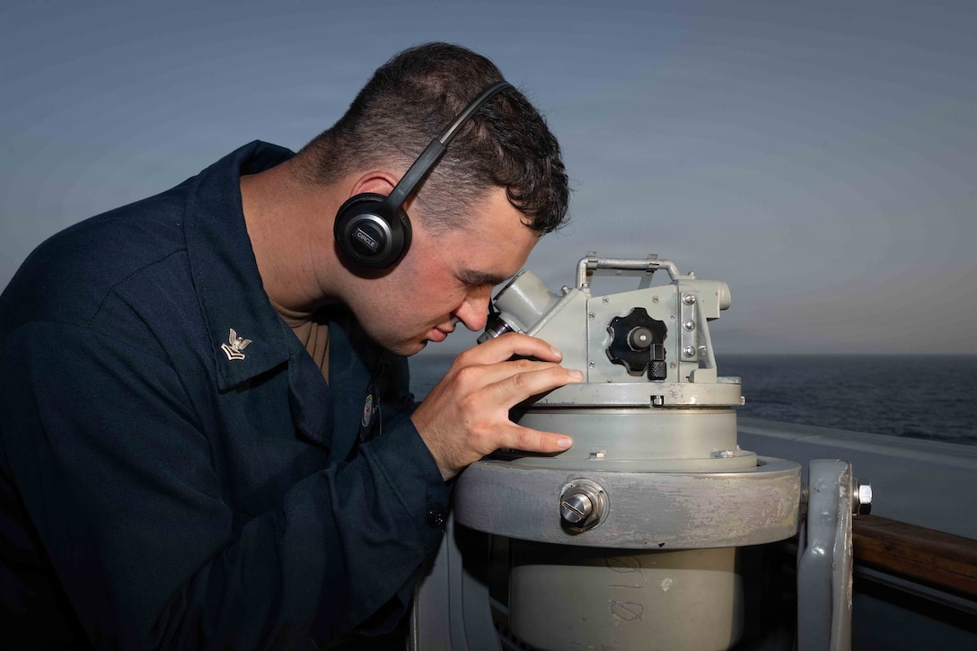 A person in a navy blue uniform and wearing headphones looks through a telescope-like instrument, with the sea and horizon in the background.