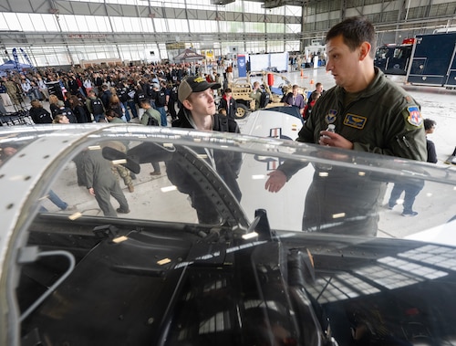 Two people talk. In front of them is an aircraft's glass canopy. Below and behind them is a large room with a large group of people.