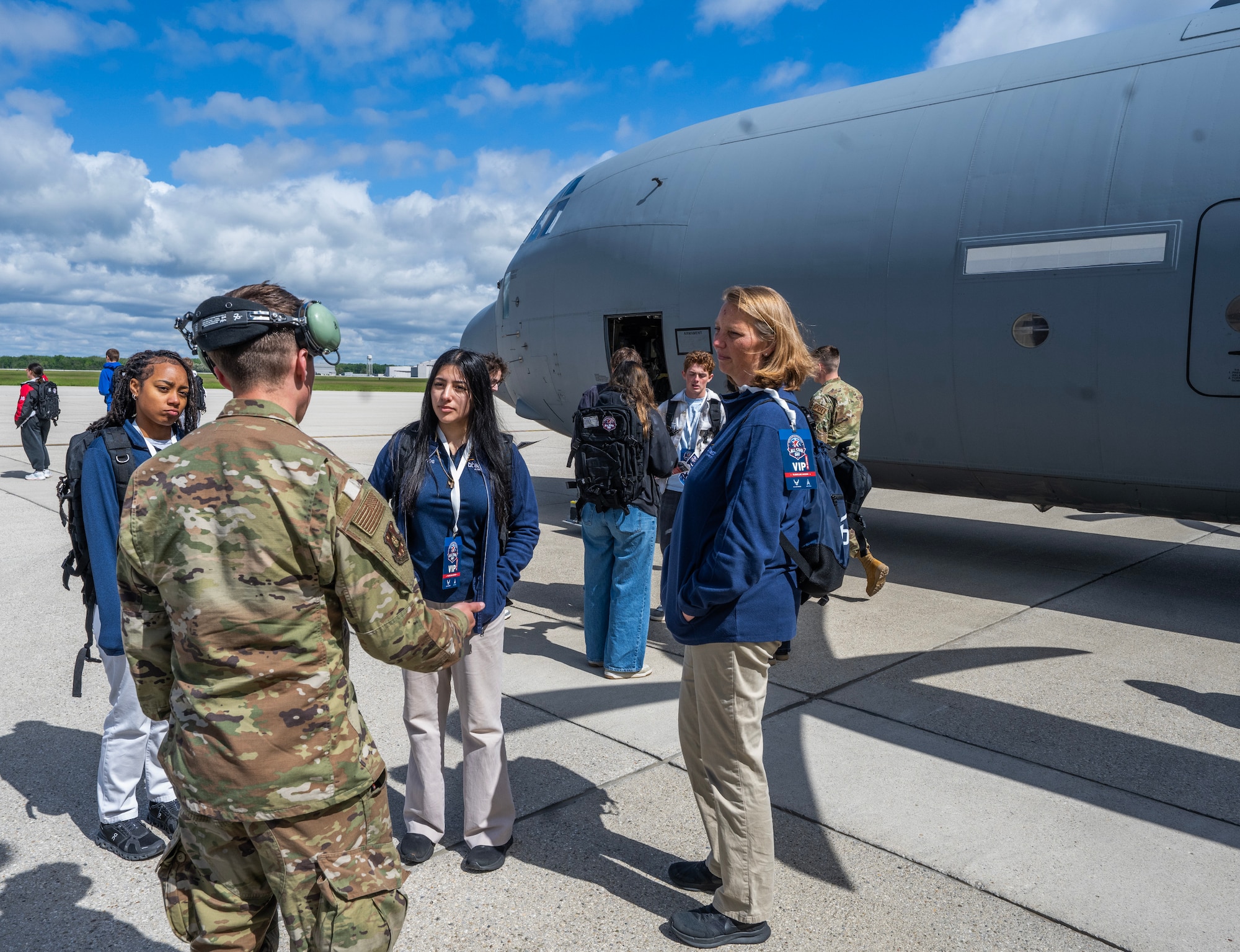A man in camouflage uniform, back to camera, talks to a couple of teenage girls and a woman. In the background is a large cargo plane.