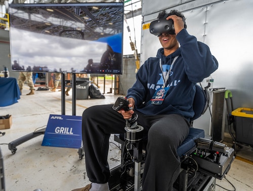 A teenage boy sits in a chair, an aircraft control stick in front of him, other controls beside him, wears VR googles as he "flys" the plane. A large monitor beside him show. what he is seeing.