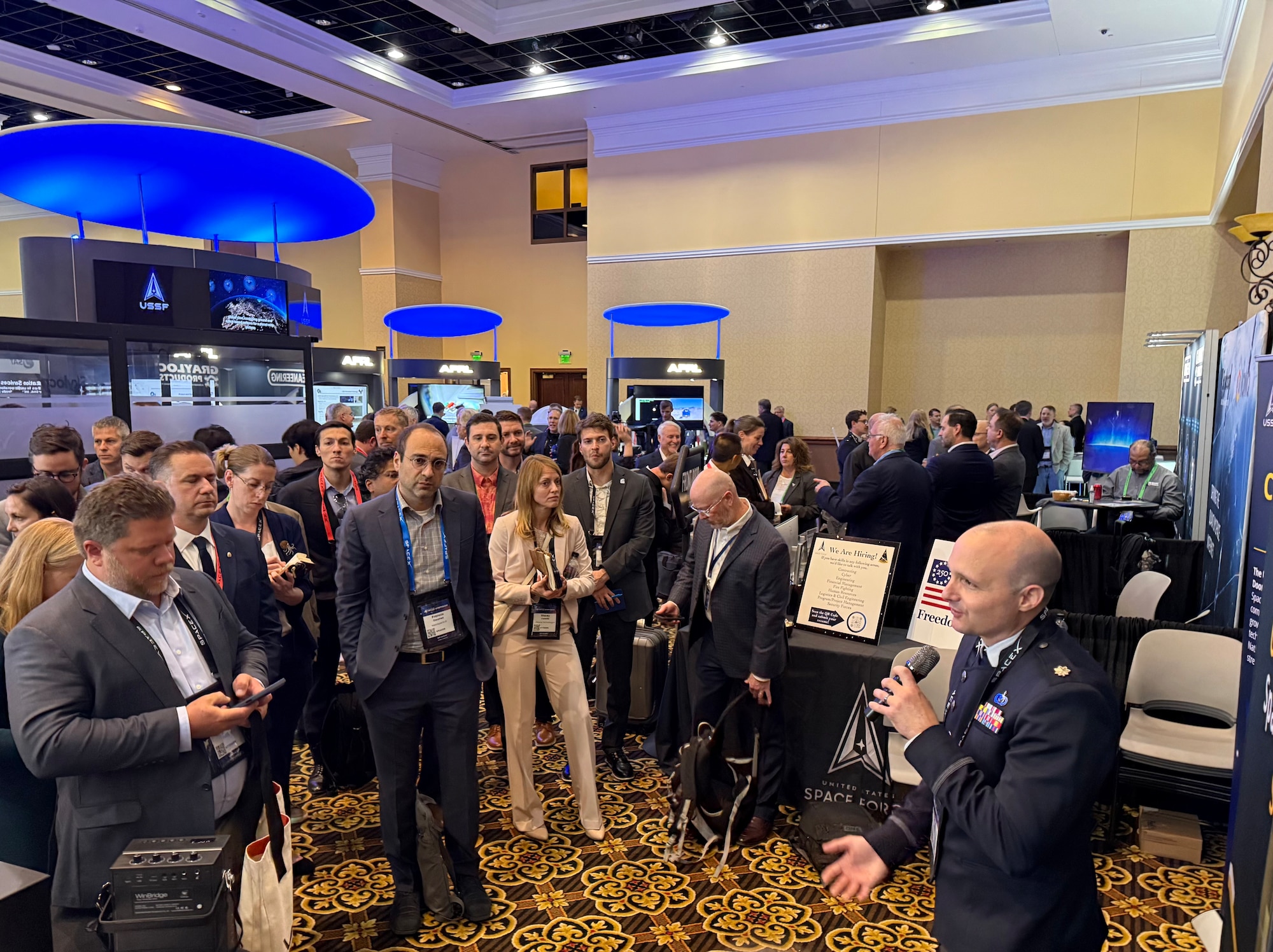 Lt. Col. Collin Greiser, System Program Manager for Space Systems Command (SSC) Advanced Battle Management, (right) addresses a crowd on the convention floor on April 15, 2026 during the 41st Space Symposium in Colorado Springs, Colorado.  Lt. Col. Greiser was a featured speaker at the United States Space Force (USSF) Front Door’s exhibit booth where he discussed the Space Domain Awareness (SDA) Tools, Applications and Processing (TAP) Lab, which he oversees.  (USSF photo by Paul Hewitt)