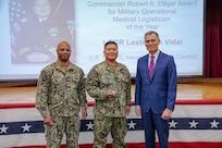 260504-N-KC192-1026 PORTSMOUTH, Va. (May 4, 2026) Cmdr. Lester Vidal, center, assigned to the Mercy-class hospital ship USNS Comfort (T-AH 20), is presented the Commander Robert A. Edgar Award for Military Operational Medical Logistician of the Year by Dr. Michael McGinnis, Bureau of Medicine and Surgery (BUMED) executive director, during the 2026 Medical Logistics Symposium held on board Naval Medical Center Portsmouth, May 4, 2026. Vidal was recognized for leading large-scale medical logistics operations and delivering critical supplies during global missions, directly enabling sustained patient care and operational reach in expeditionary environments. The Navy Medicine Logistics Symposium aims to maintain a common operating picture and update the Medical Logistics community regarding the fiscal year 2026 Navy Medicine Campaign Order and the Navy MEDLOG goal to build a logistics system that delivers ready operational units to support the Naval Strategy. (U.S. Navy photo by Mass Communication Specialist Second Class Levi Decker)