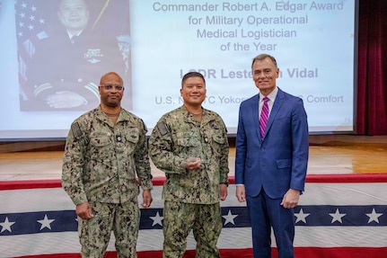 260504-N-KC192-1026 PORTSMOUTH, Va. (May 4, 2026) Cmdr. Lester Vidal, center, assigned to the Mercy-class hospital ship USNS Comfort (T-AH 20), is presented the Commander Robert A. Edgar Award for Military Operational Medical Logistician of the Year by Dr. Michael McGinnis, Bureau of Medicine and Surgery (BUMED) executive director, during the 2026 Medical Logistics Symposium held on board Naval Medical Center Portsmouth, May 4, 2026. Vidal was recognized for leading large-scale medical logistics operations and delivering critical supplies during global missions, directly enabling sustained patient care and operational reach in expeditionary environments. The Navy Medicine Logistics Symposium aims to maintain a common operating picture and update the Medical Logistics community regarding the fiscal year 2026 Navy Medicine Campaign Order and the Navy MEDLOG goal to build a logistics system that delivers ready operational units to support the Naval Strategy. (U.S. Navy photo by Mass Communication Specialist Second Class Levi Decker)