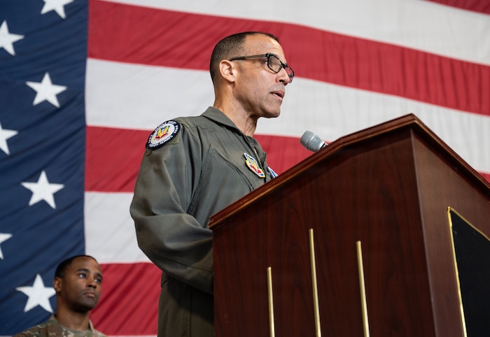 An Air Force pilot speaks a podium with a giant American flag in the background