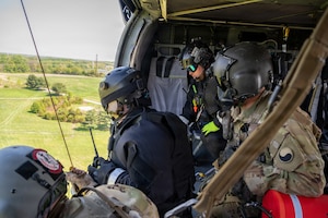 Two service members in camouflage uniforms and two other people in black flight suits sit inside a flying helicopter.