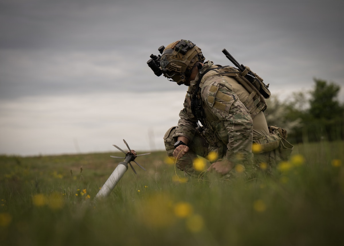 an eod member crouched in a green field with yellow flowers, a uxo to his left