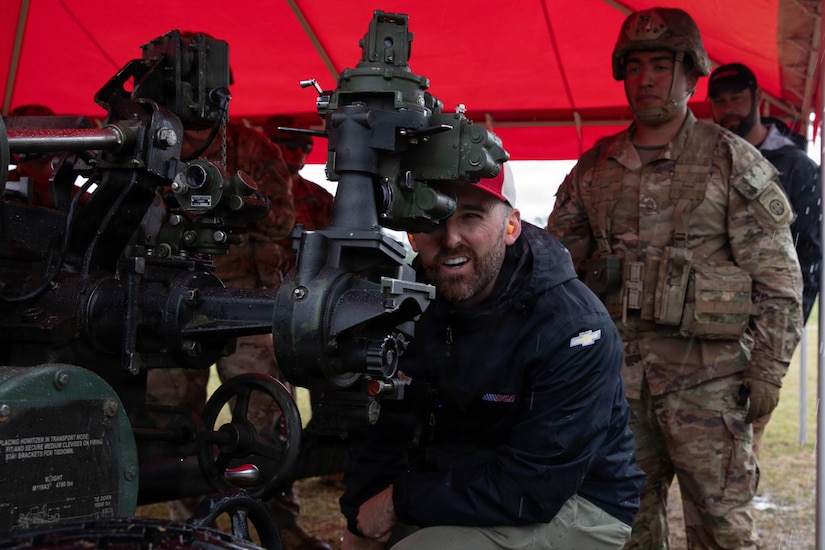 A man wearing a red baseball cap and blue jacket looks through the sight of a Howitzer. Two men in camouflage military uniforms stand behind him.