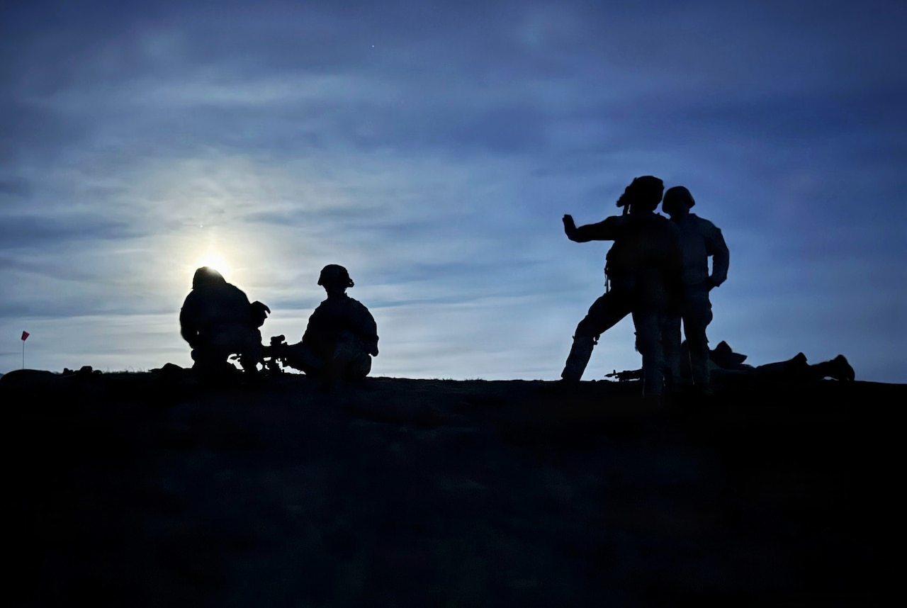 A silhouette of five people wearing helmets standing, sitting and lying down on a hill; two of the people are holding rifles.