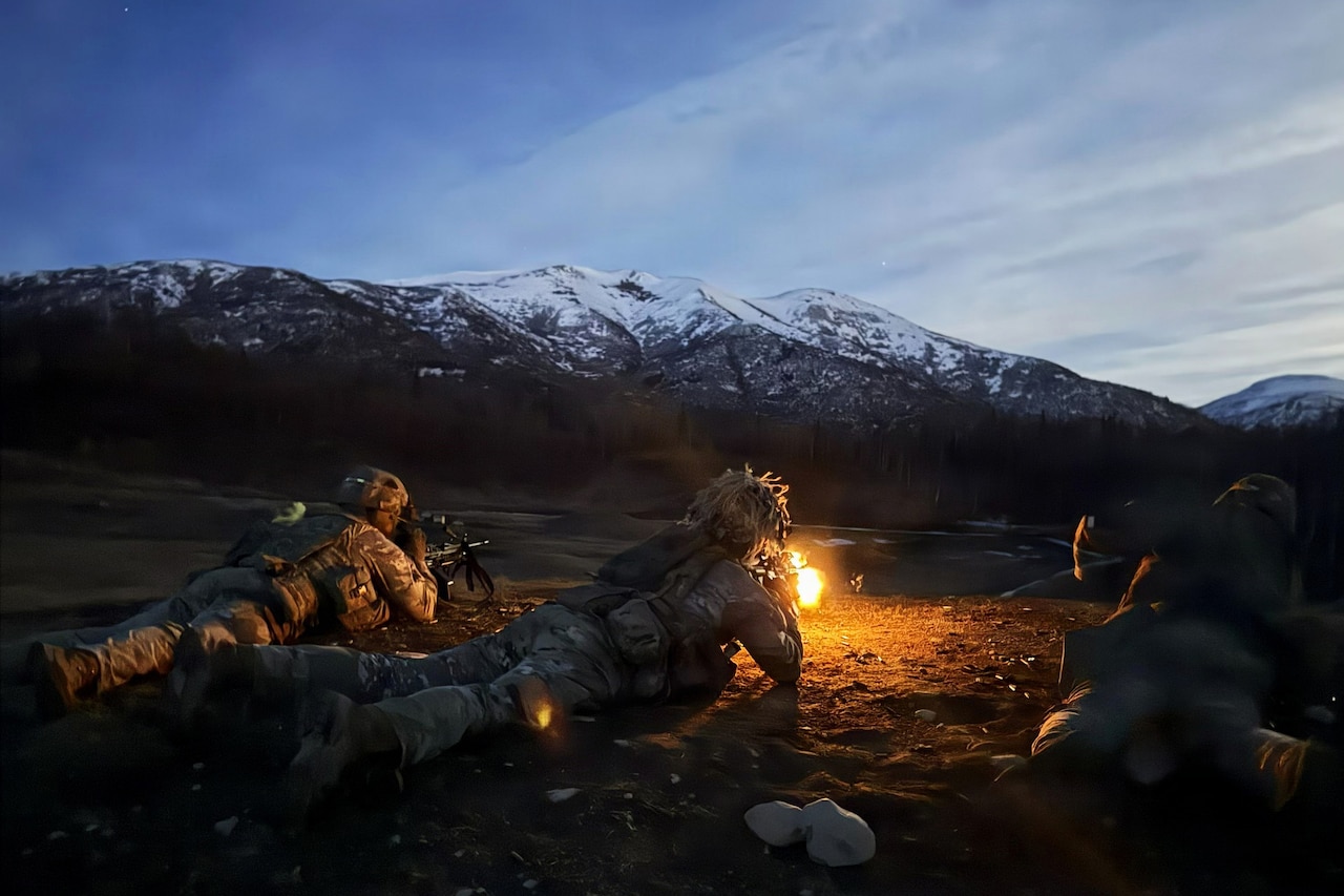 Two people in camouflage military uniforms lie on the ground, firing rifles into an open field, while another man in similar attire lies next to them, observing; there are trees and mountains in the background.