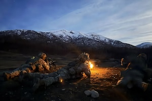 Two people in camouflage military uniforms lie on the ground, firing rifles into an open field, while another man in similar attire lies next to them, observing; there are trees and mountains in the background.