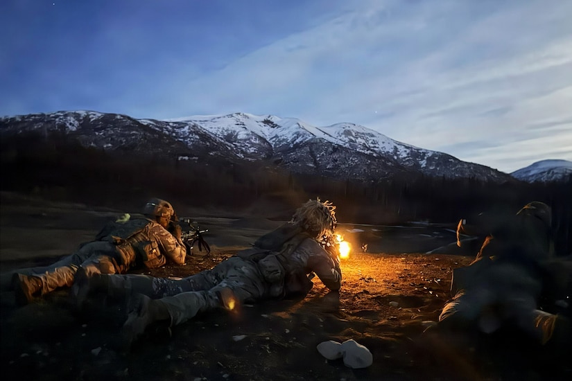 Two people in camouflage military uniforms lie on the ground, firing rifles into an open field, while another man in similar attire lies next to them, observing; there are trees and mountains in the background.