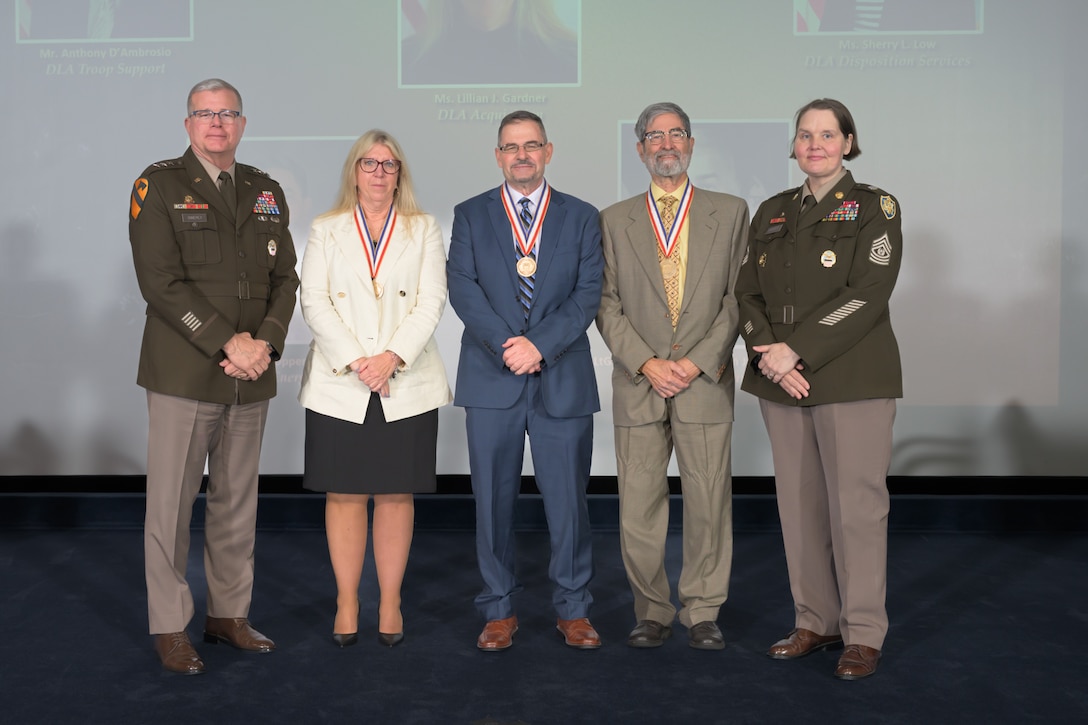 Three hall of fame inductees are posed facing the camera on a stage flanked by the DLA director and senior enlisted leader.