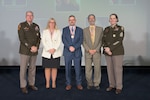 Three hall of fame inductees are posed facing the camera on a stage flanked by the DLA director and senior enlisted leader.
