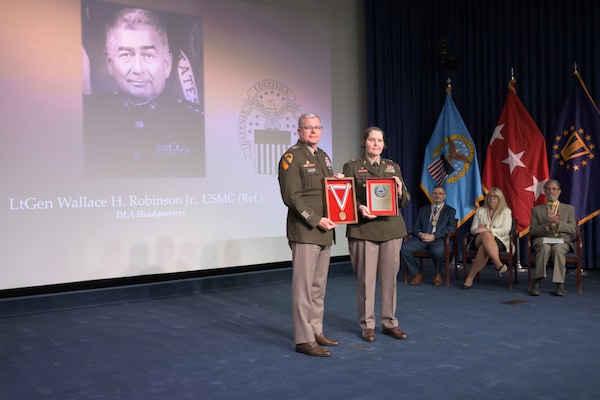 Lt. Gen. Mark Simerly and Sgt. Maj. Petra Casarez hold a medal and plaque with Lt. Gen. Wallace Robinson Jr.'s portrait projected behind them on stage, with other Hall of Fame inductees seated to the side of the stage.