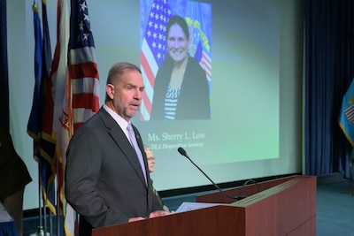 Mike Cannon speaks into a microphone on stage at a podium, with flags and a projected picture of Sherry Low on the screen behind him.