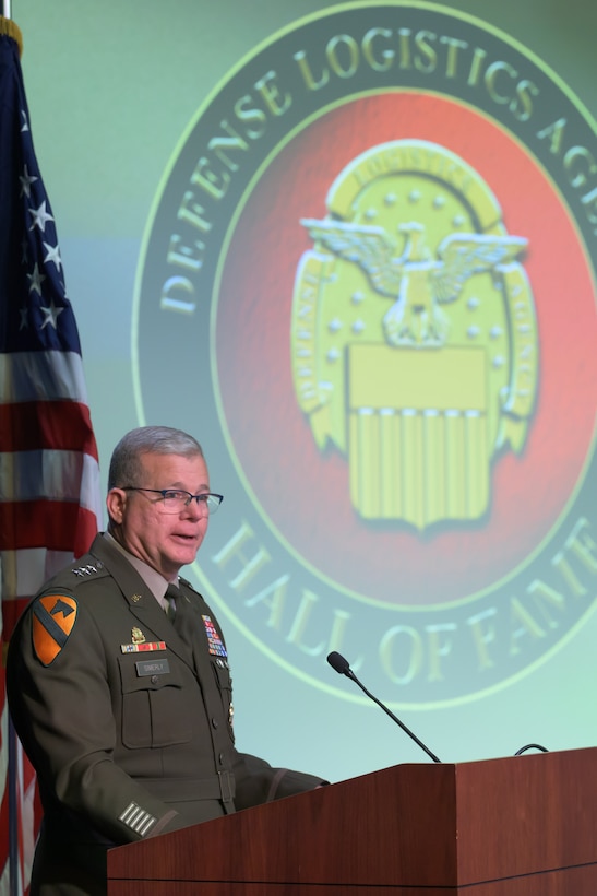 Lt. Gen. Mark Simerly speaks from behind a podium with a microphone, with the Defense Logistics Agency Hall of Fame emblem projected on the screen over his shoulder.