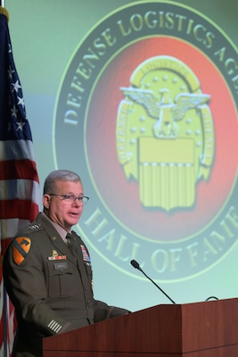 Lt. Gen. Mark Simerly speaks from behind a podium with a microphone, with the Defense Logistics Agency Hall of Fame emblem projected on the screen over his shoulder.