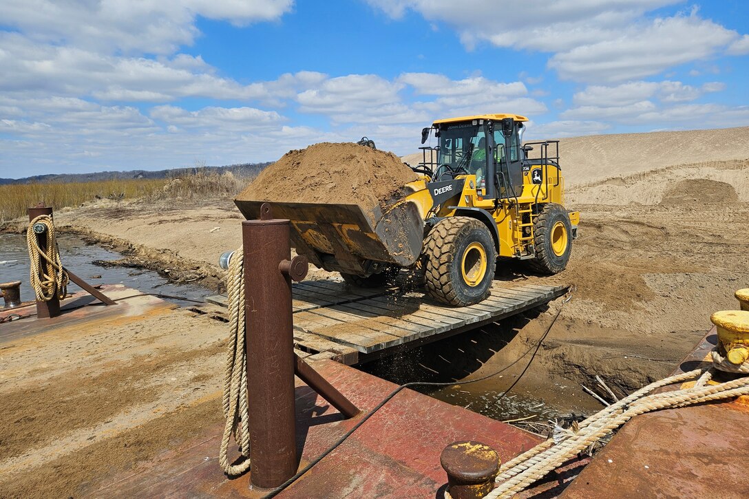A loader carries sand from a barge into a dredge material placement site along the river.