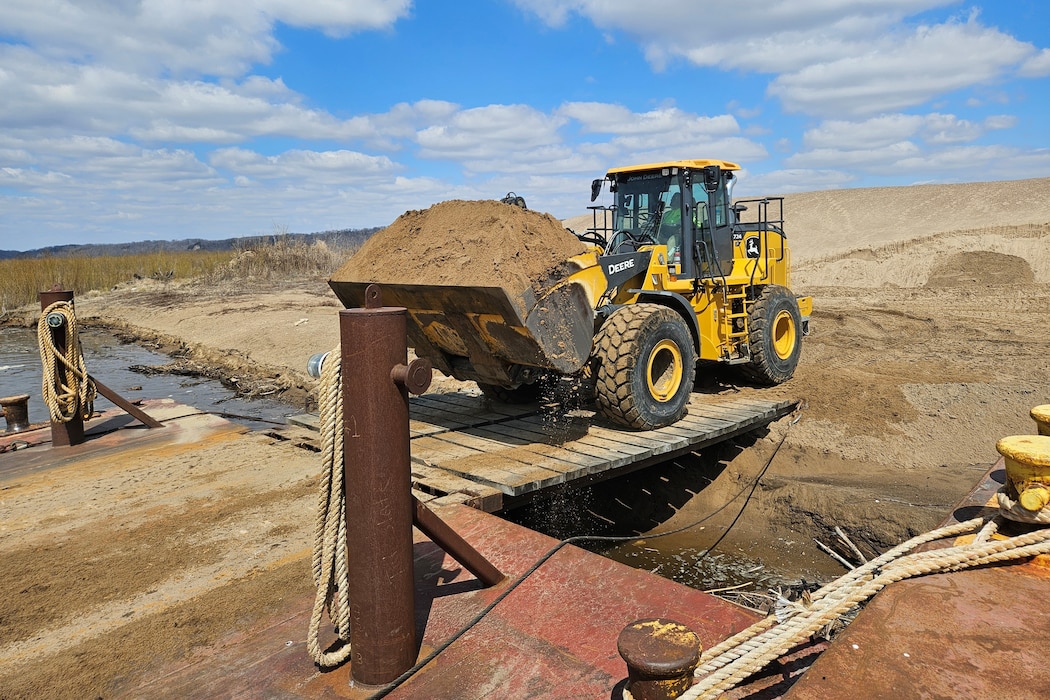 A loader carries sand from a barge into a dredge material placement site along the river.