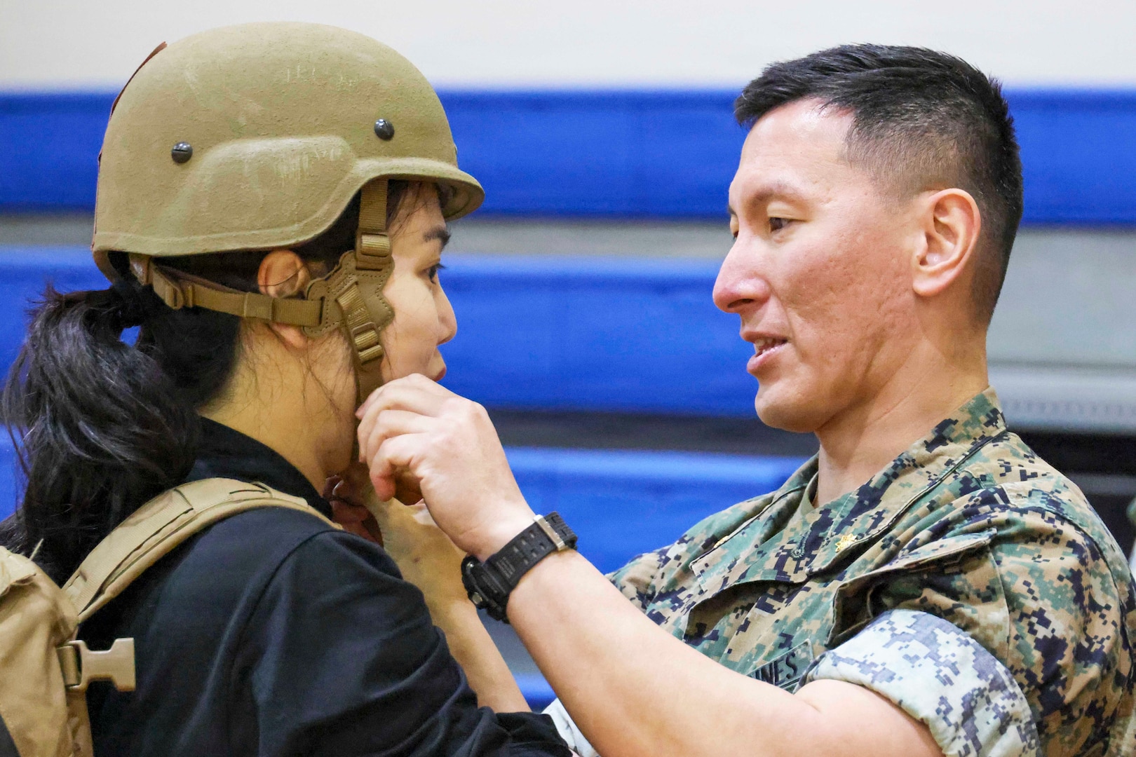 A Marine faces his wife while adjusting a helmet on her head in front of blue benches.