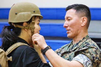 A Marine faces his wife while adjusting a helmet on her head in front of blue benches.