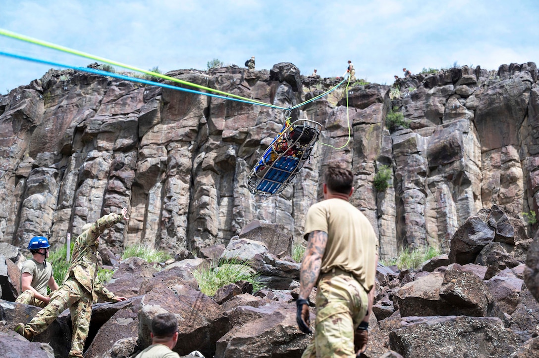 Airmen at the bottom of a cliff watch as service members at the top of the cliff pull a basket stretcher with blue and green ropes.