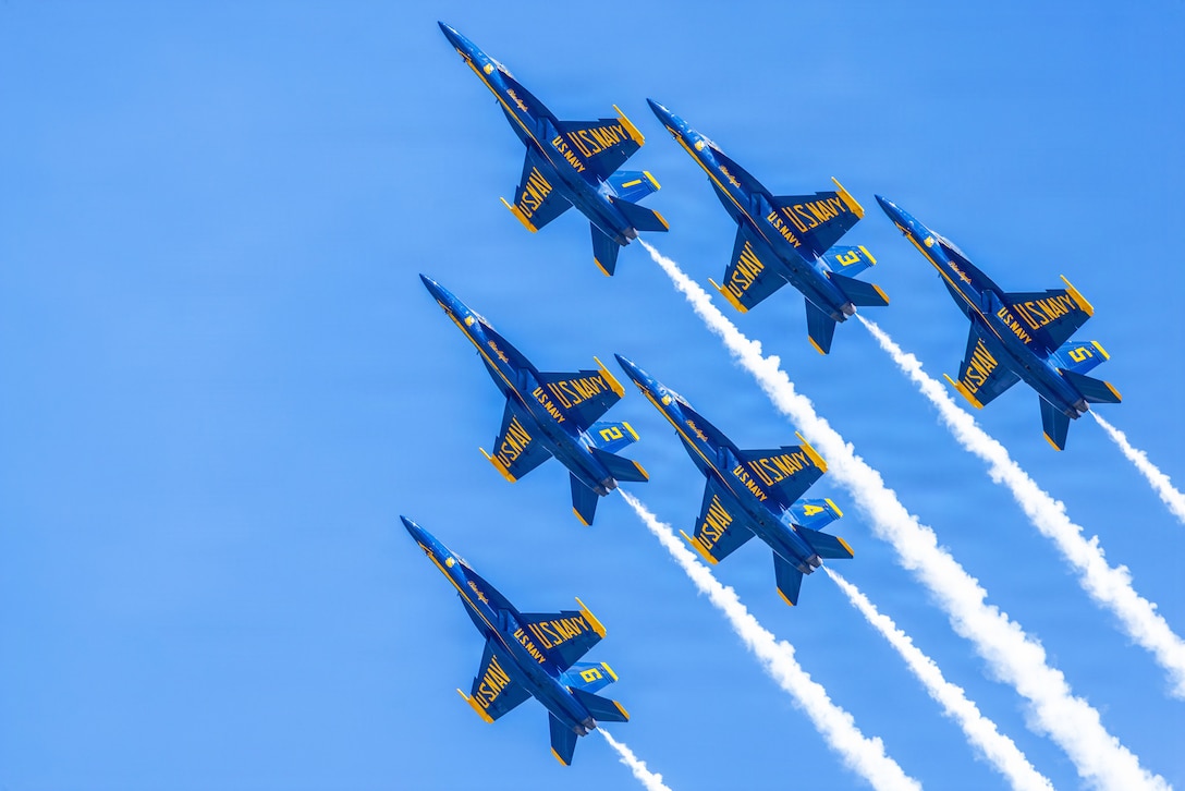 Six blue and yellow aircraft ascend diagonally in formation in a blue sky, leaving behind white trails of smoke.