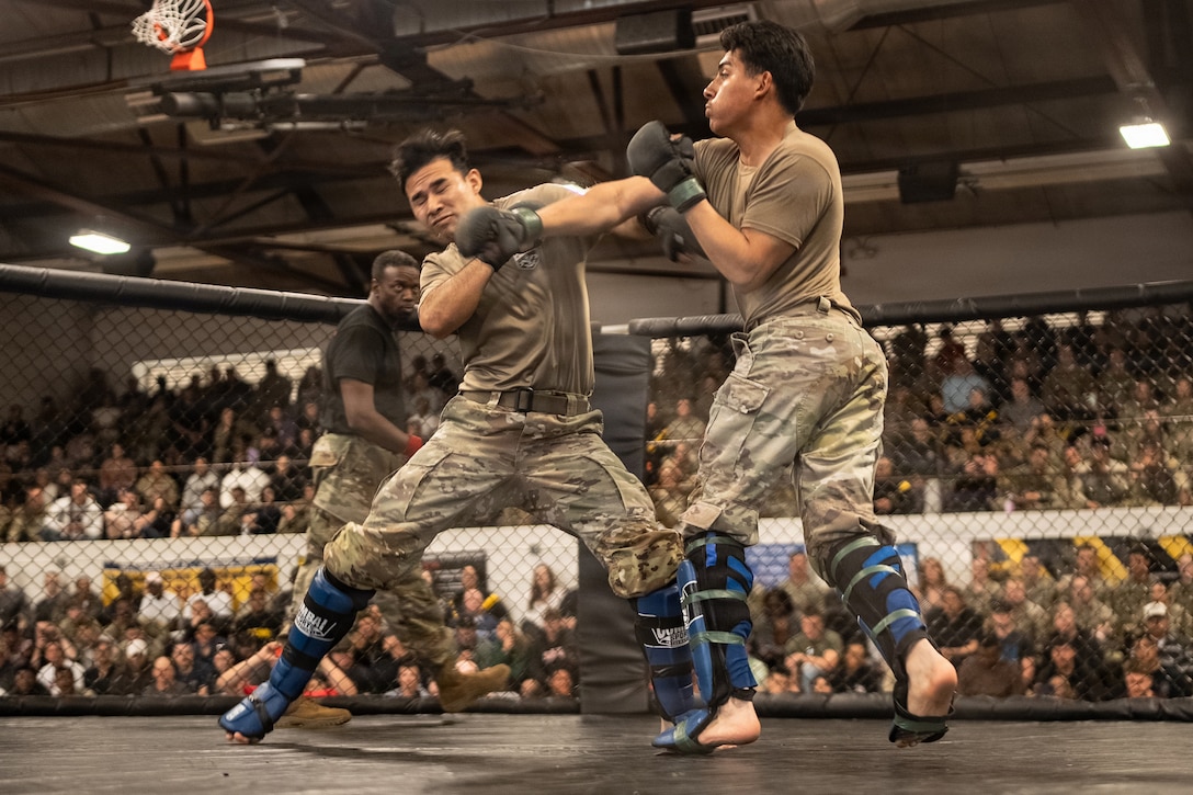 Two soldiers wearing boxing gloves and leg and toe pads fight in a fenced-in ring, with a referee and people in stands in the background of an auditorium.
