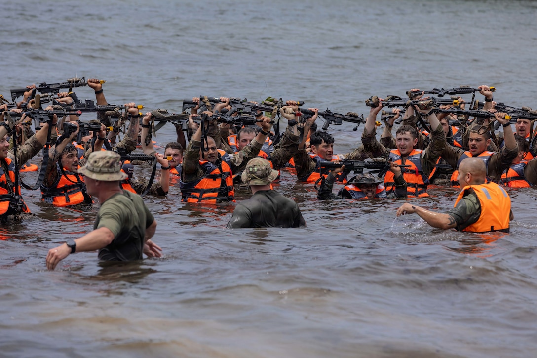 Soldiers in orange vests raise their arms above their heads while standing in the ocean, with the ends of rifles in their hands. Three instructors face them in the water.