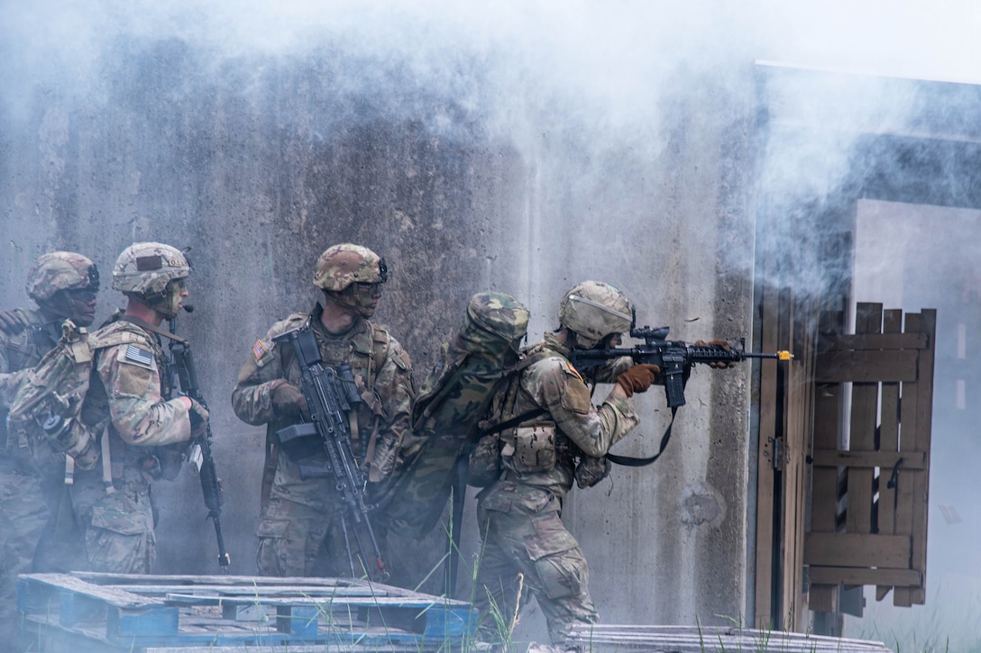 A soldier points a machine gun ahead, with part of a wooden gate in front of him, three soldiers standing behind him holding machine guns and smoke around them.
