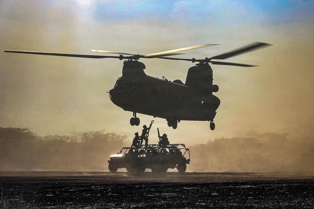 Two soldiers stand on vehicles and point slings toward the bottom of a helicopter hovering above them, with another soldier kneeling to support one of the soldiers. They are all in silhouette.