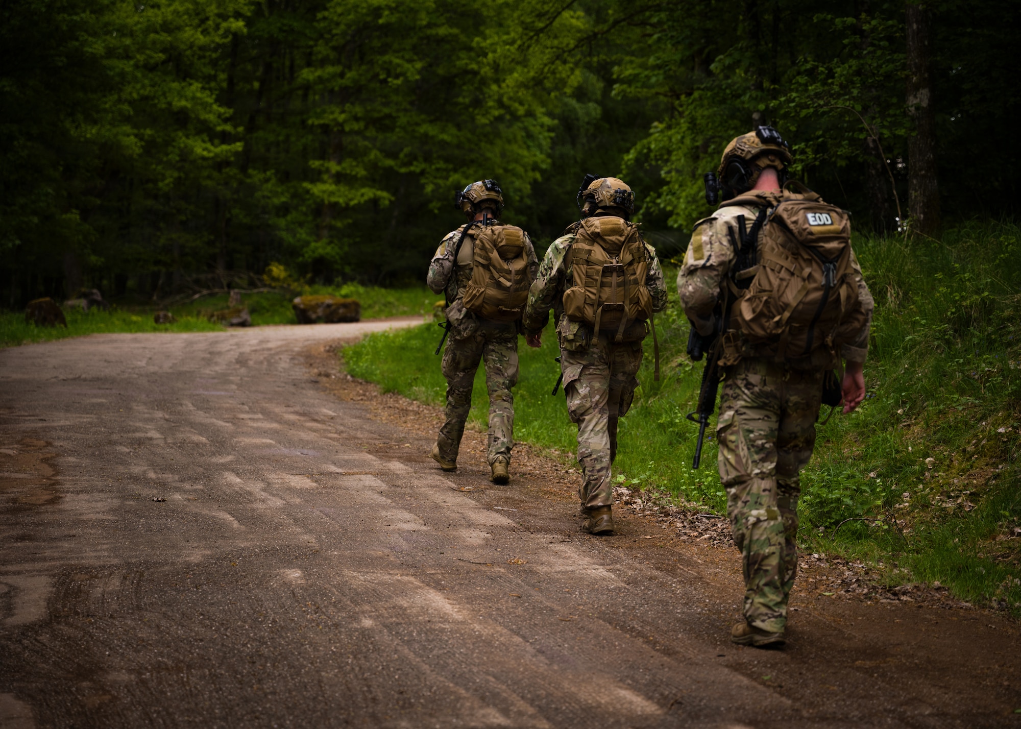 three eod members, backs turned from camera, walking on a dirt path through a forest