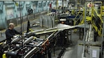 Workers oversee artillery shell production inside a munitions manufacturing facility. Rows of large shell casings move through an industrial assembly line surrounded by heavy machinery, conveyor systems, and production equipment.