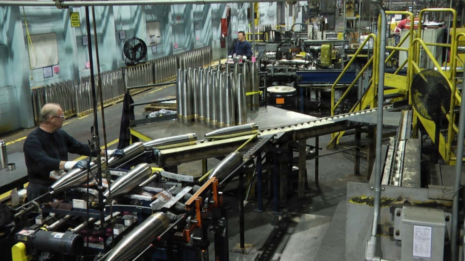 Workers oversee artillery shell production inside a munitions manufacturing facility. Rows of large shell casings move through an industrial assembly line surrounded by heavy machinery, conveyor systems, and production equipment.