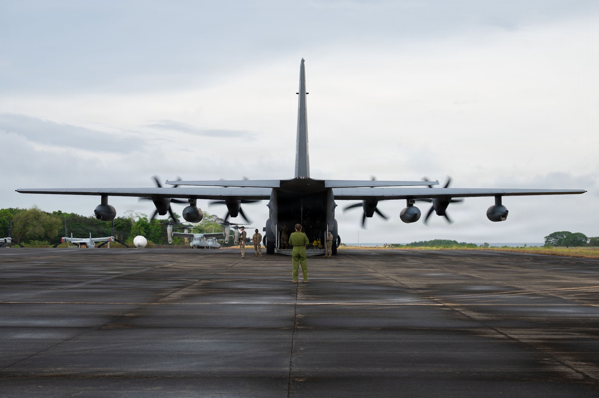 A flight nurse, viewed from behind, directs medical personnel toward the open rear ramp of a large military cargo aircraft. The aircraft sits center-frame, with its wings stretching outward to create striking, symmetrical leading lines across the image.