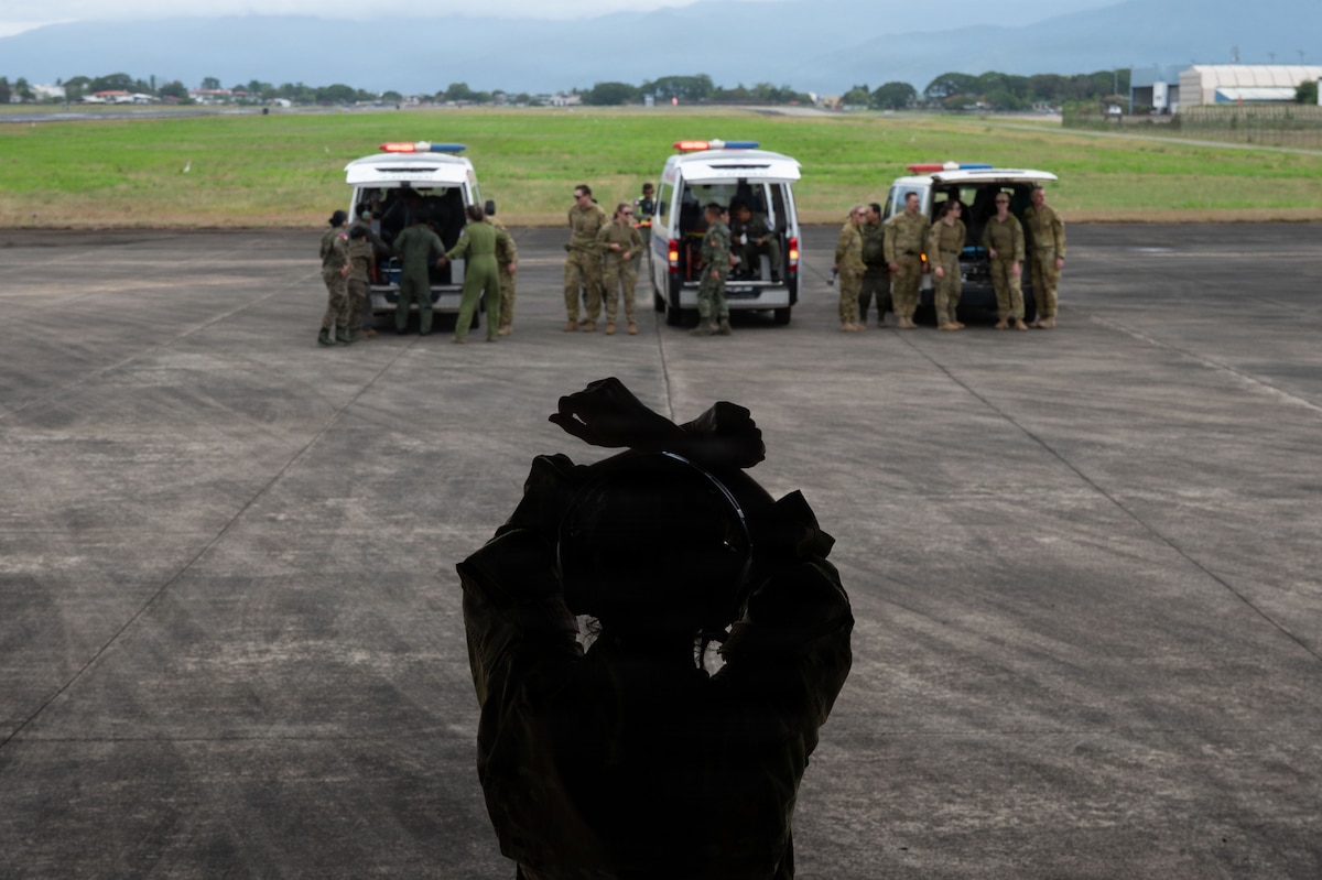 A silhouetted flight nurse, seen from behind, uses hand signals to direct medical personnel gathered around three ambulances parked on the airfield. Mountains are visible in the distance.