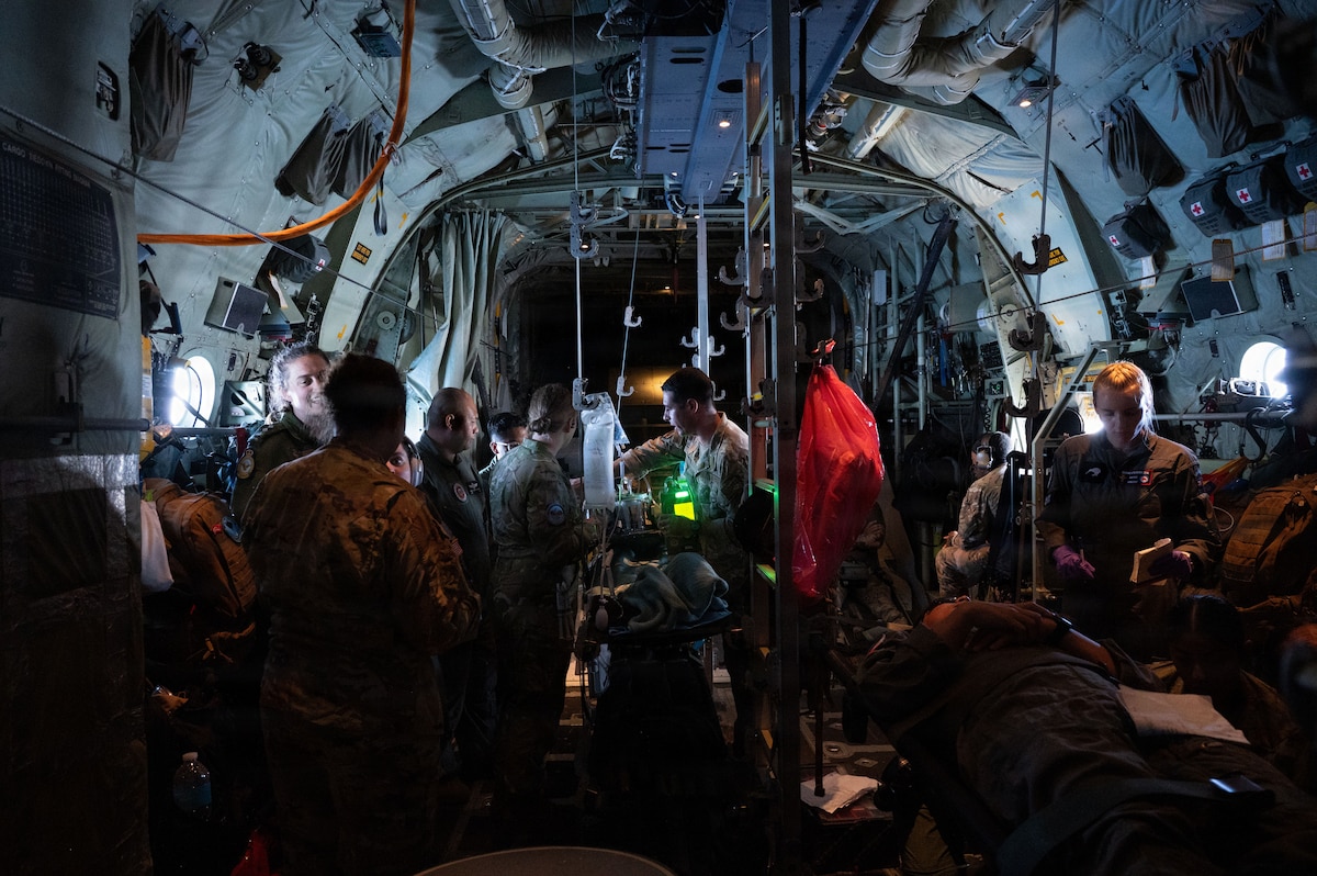 Inside the busy interior of a military aircraft, a diverse group of medical personnel in various uniforms work together around a simulated patient on a litter. The scene is crowded with service members and equipment, showing a realistic in-flight training scenario.