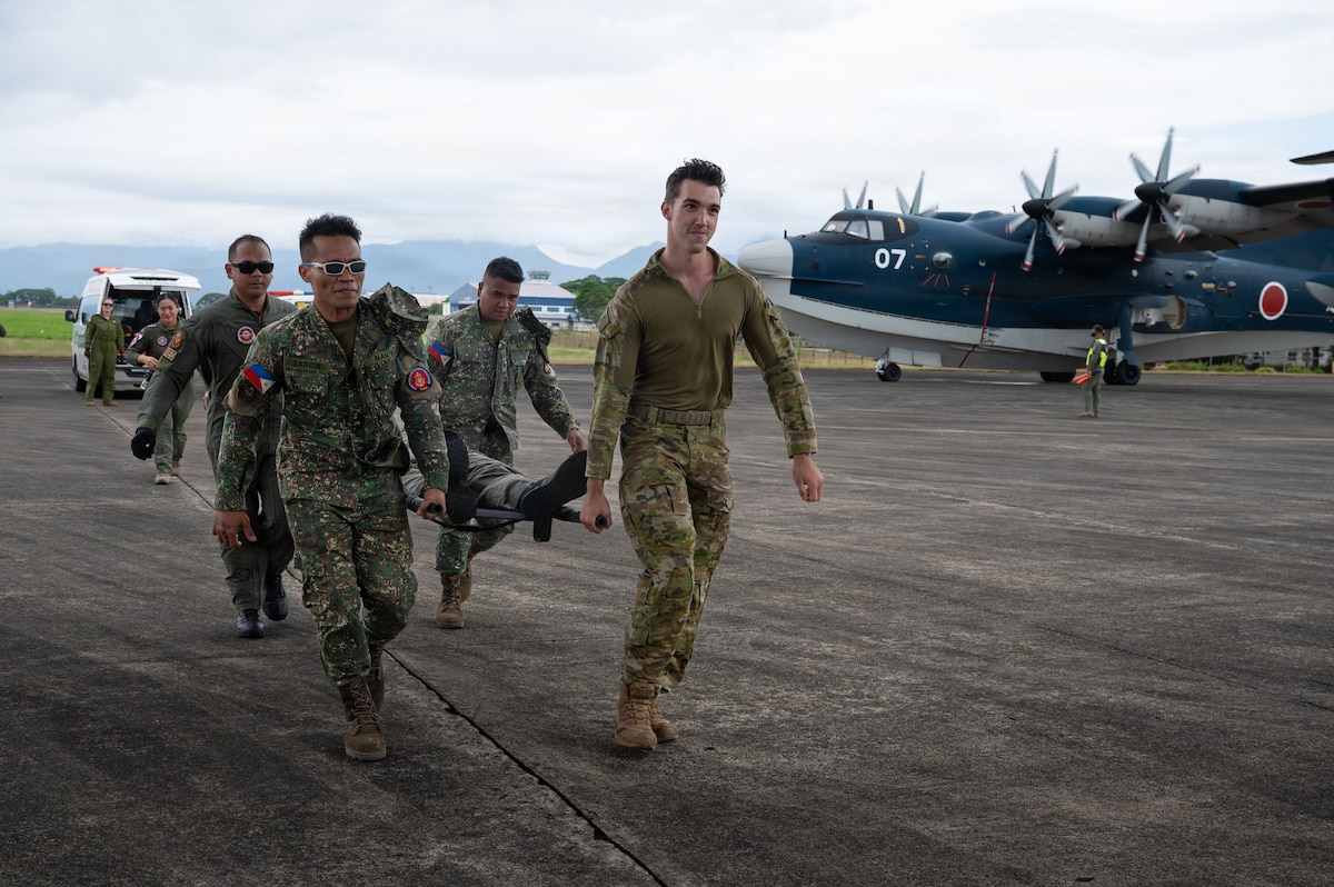 A multinational team of service members carries a patient on a litter across an airfield. A large, blue and white multi-propeller aircraft and an ambulance are visible in the background.