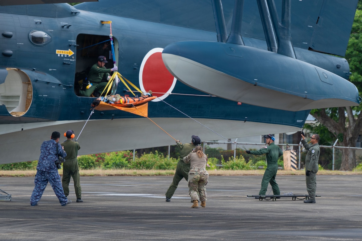 A multinational team of medical personnel works together to lower a patient on a stretcher from the side door of a large, blue aircraft. Using a system of ropes, the team on the ground guides the stretcher as it is passed out of the aircraft by a service member inside.