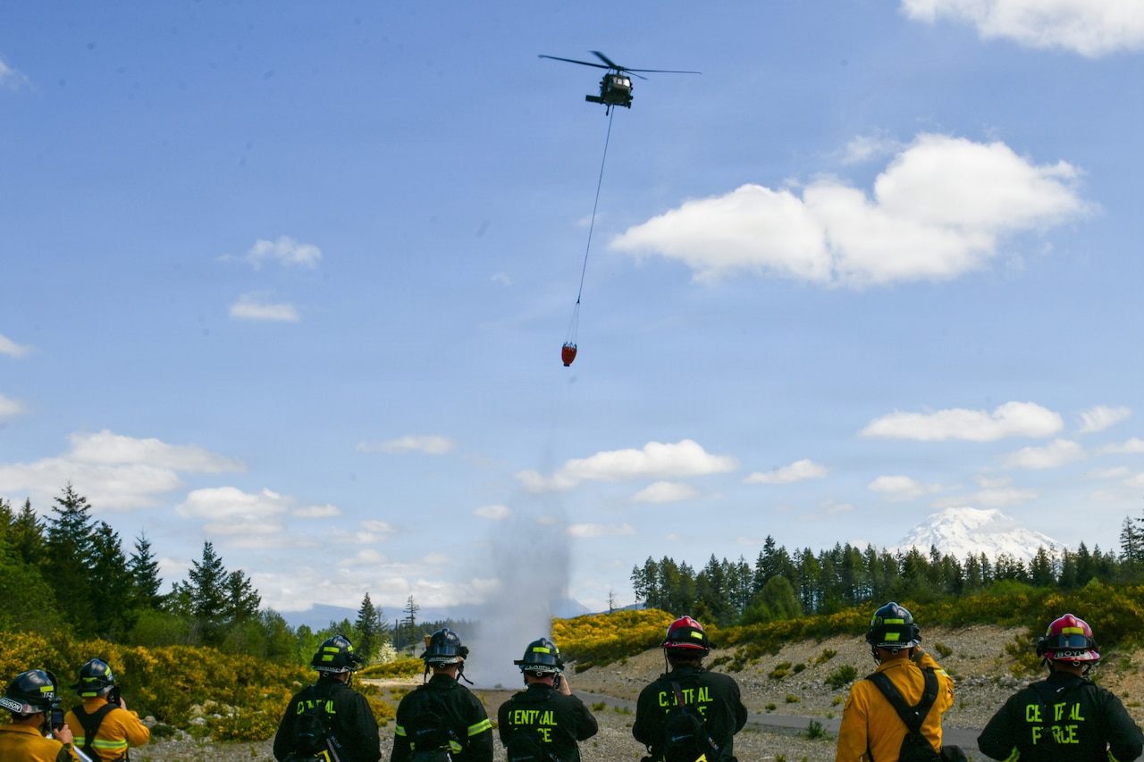 A military helicopter drops water on the ground in an open area surrounded by trees as eight people in firefighter uniforms observe in the foreground.