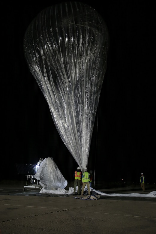 Two people in safety vests and helmets hold the ends of twine attached to a large silver balloon floating in the night sky.