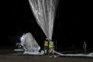 Two people in safety vests and helmets hold the ends of twine attached to a large silver balloon floating in the night sky.