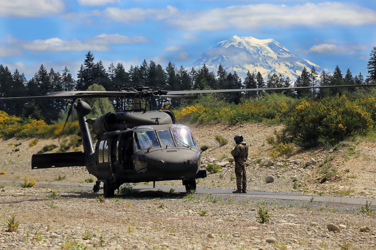 A person wearing a camouflage military uniform stands next to a military helicopter preparing to take off from a training location outside; there are trees and mountains in the background.
