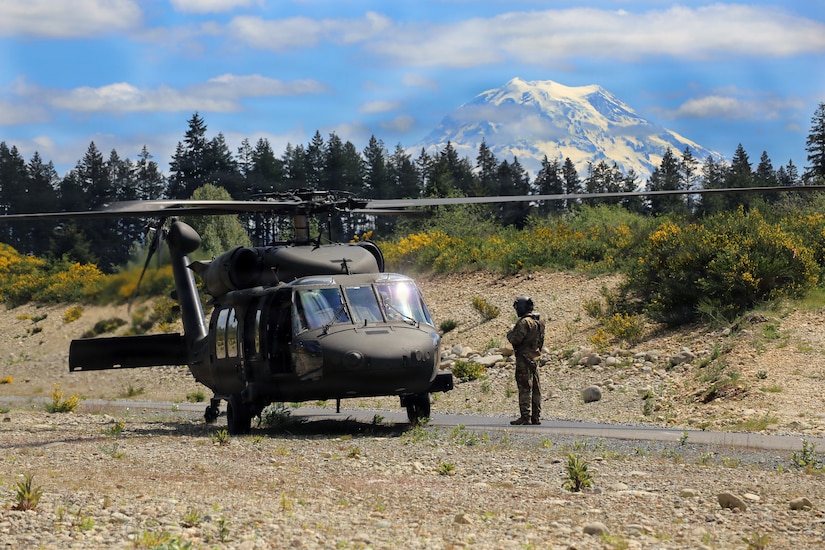 A person wearing a camouflage military uniform stands next to a military helicopter preparing to take off from a training location outside; there are trees and mountains in the background.