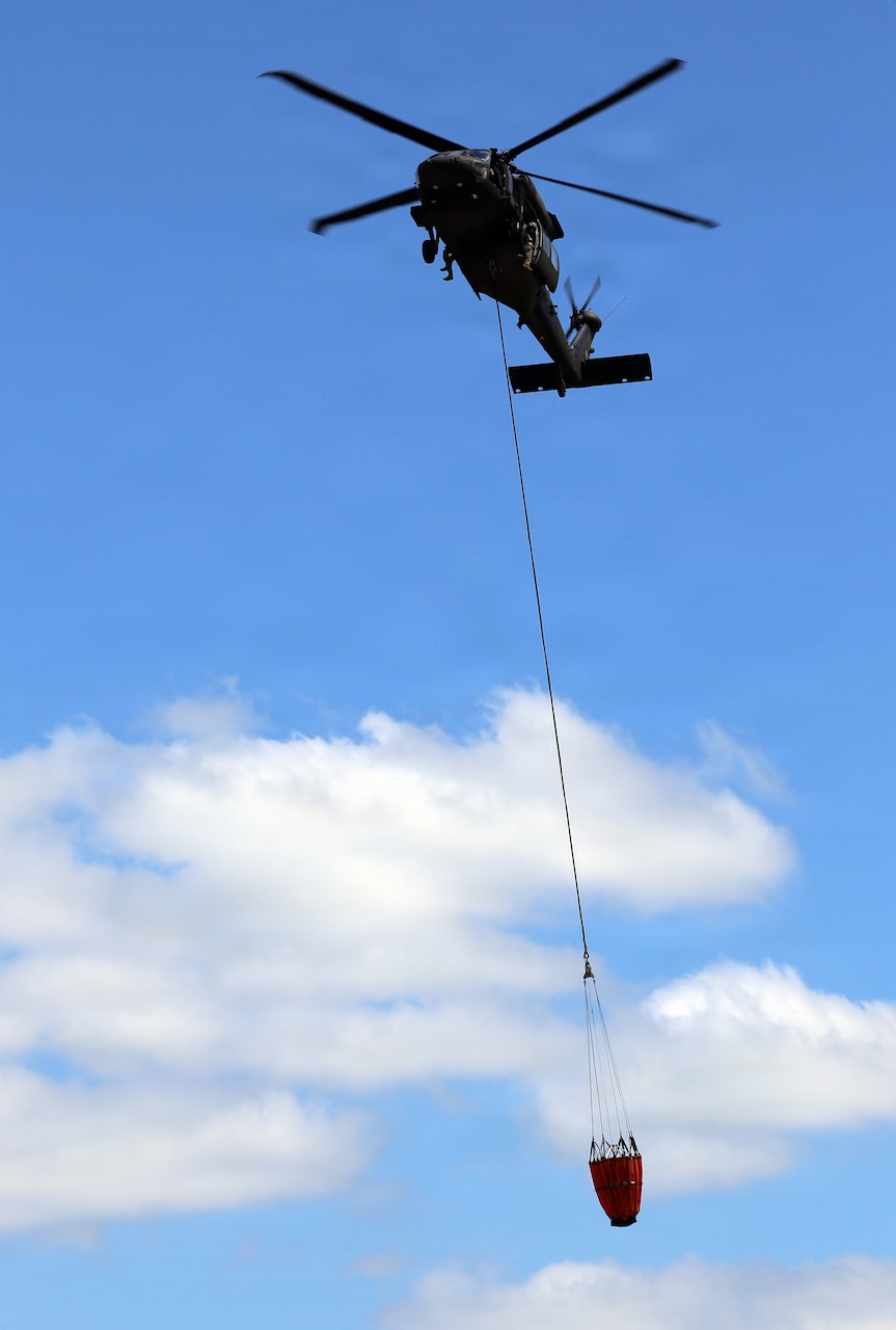 A military helicopter carries a large bucket of water that is hanging from a cable attached to the bottom of the aircraft.