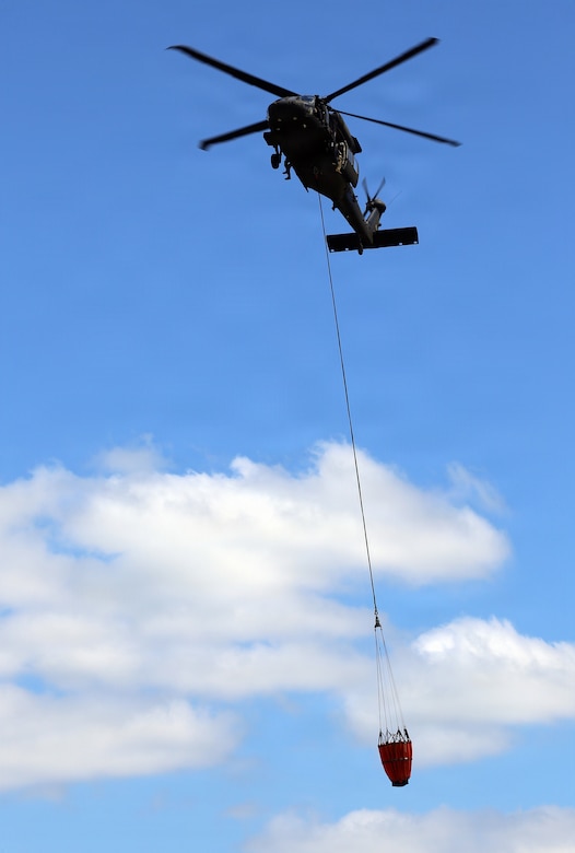 A military helicopter carries a large bucket of water that is hanging from a cable attached to the bottom of the aircraft.