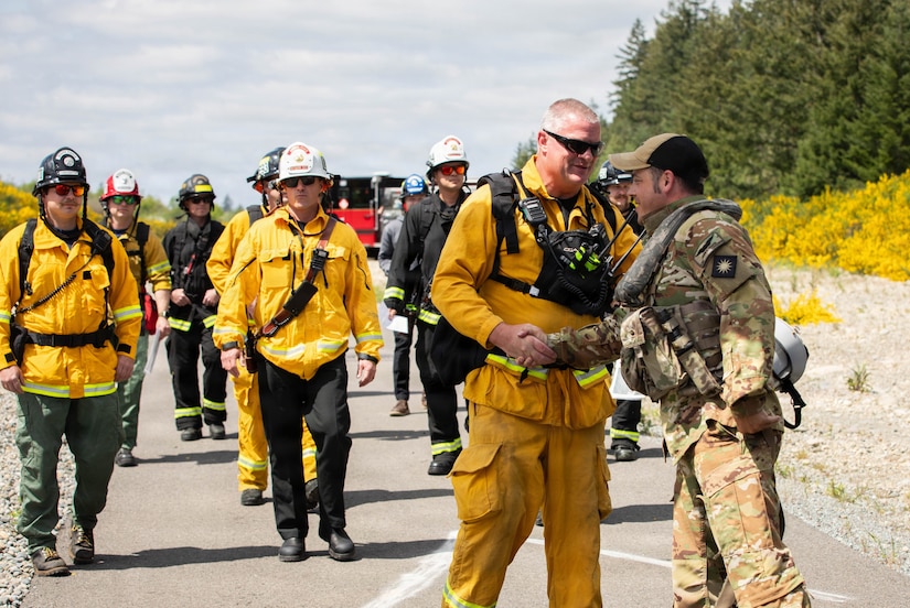 A man wearing a camouflage military uniform shakes hands with another man in a firefighter uniform at a training location outside; there are several other men wearing firefighter uniforms and helmets standing in the background.